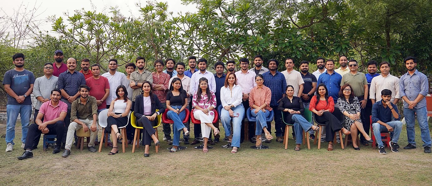 A large team seated and standing together outdoors under trees at an offsite.